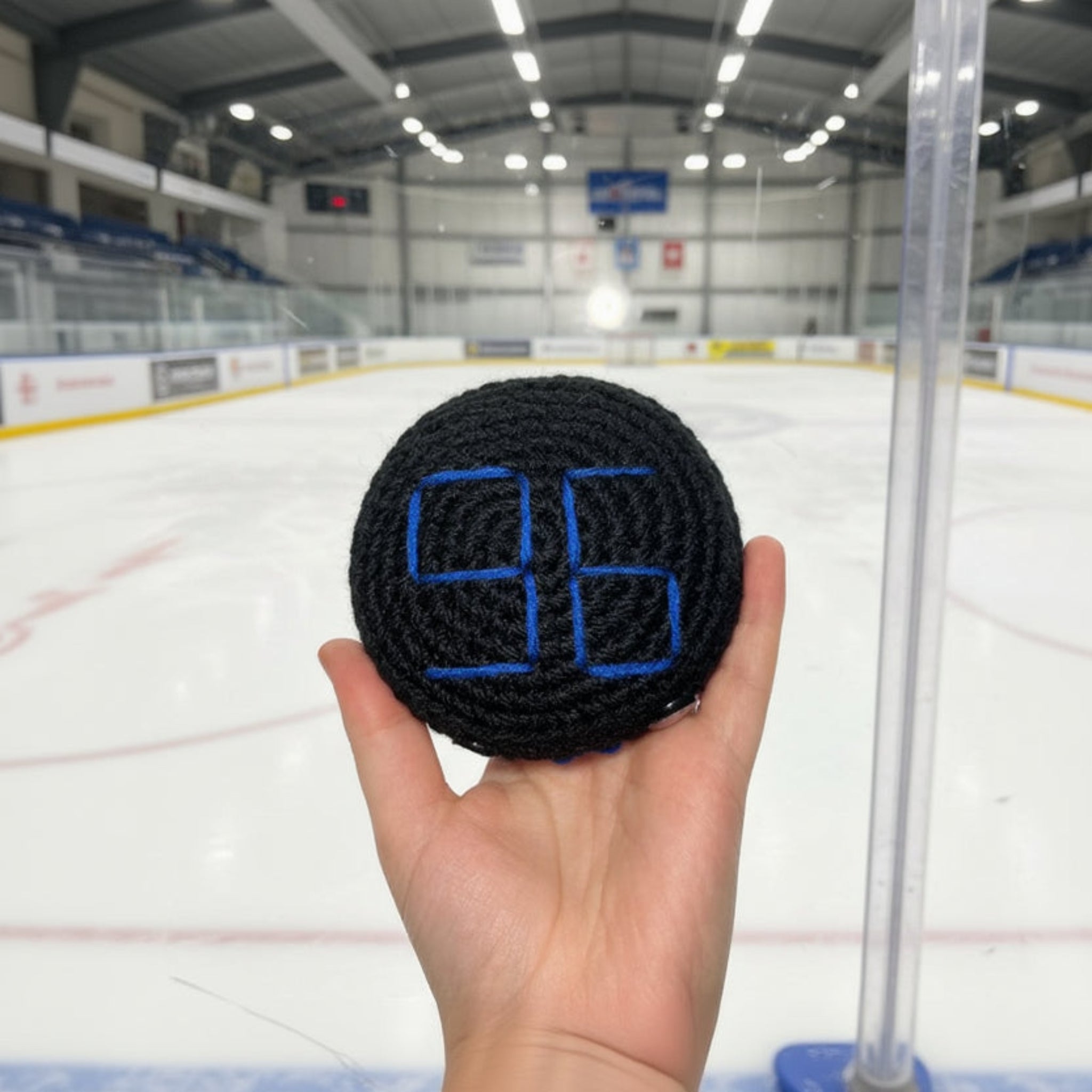 Hand holding black crochet hockey puck plush with coventry blaze blue themed colour stitching in front of an ice rink