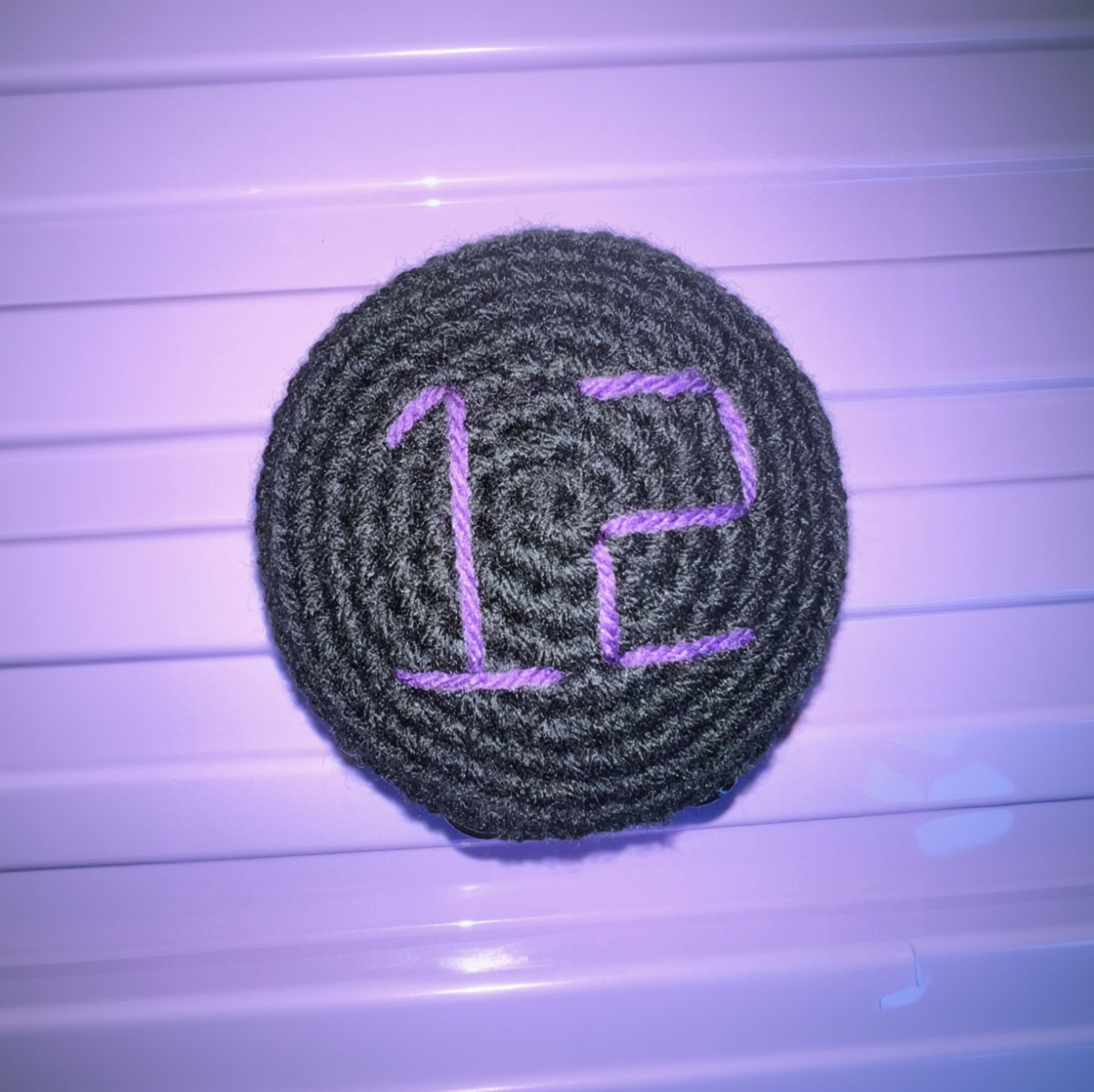 Black crochet hockey puck with glasgow clan purple stitched number on top sitting on a purple background