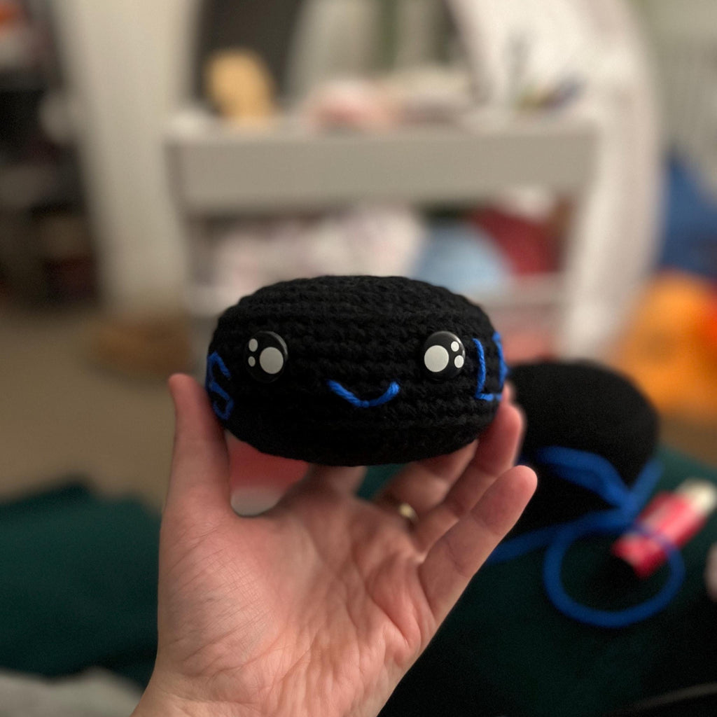 Hand holding black crochet ice hockey puck plush with blue stitching and a smiley face