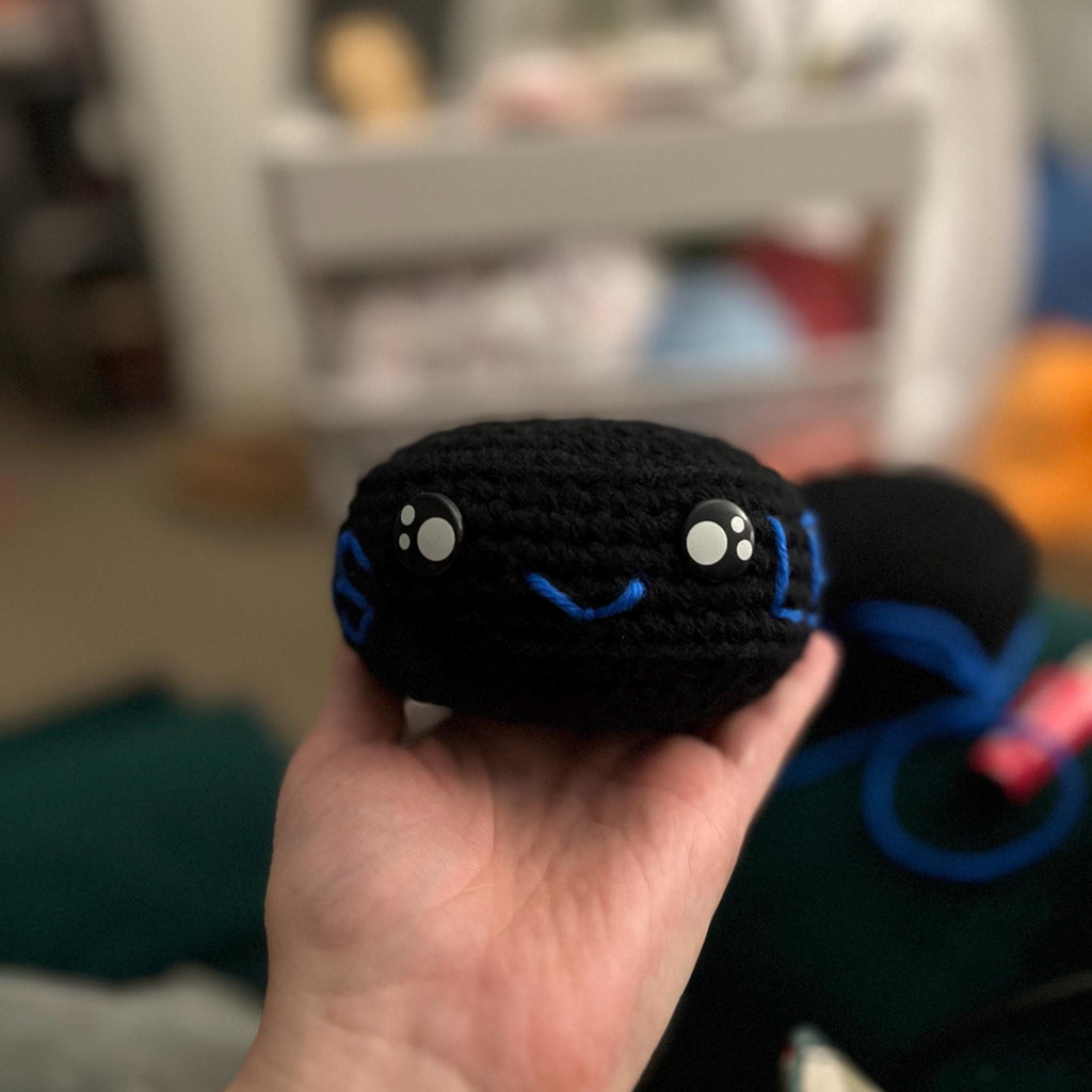 Hand holding black crochet ice hockey puck plush with blue stitching and a smiley face