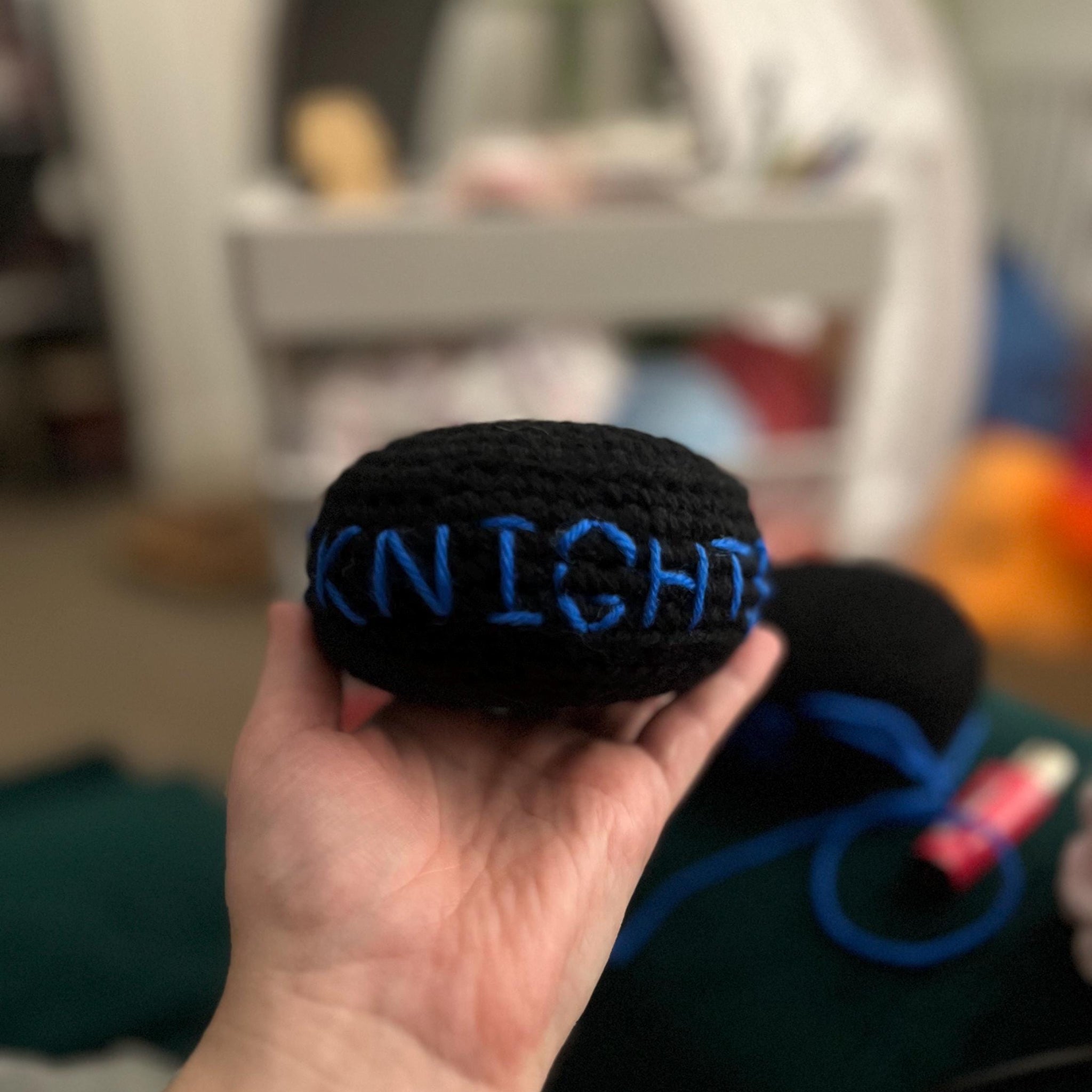 Hand holding one black crochet plush ice hockey puck with blue stitching of 'KNIGHTS'