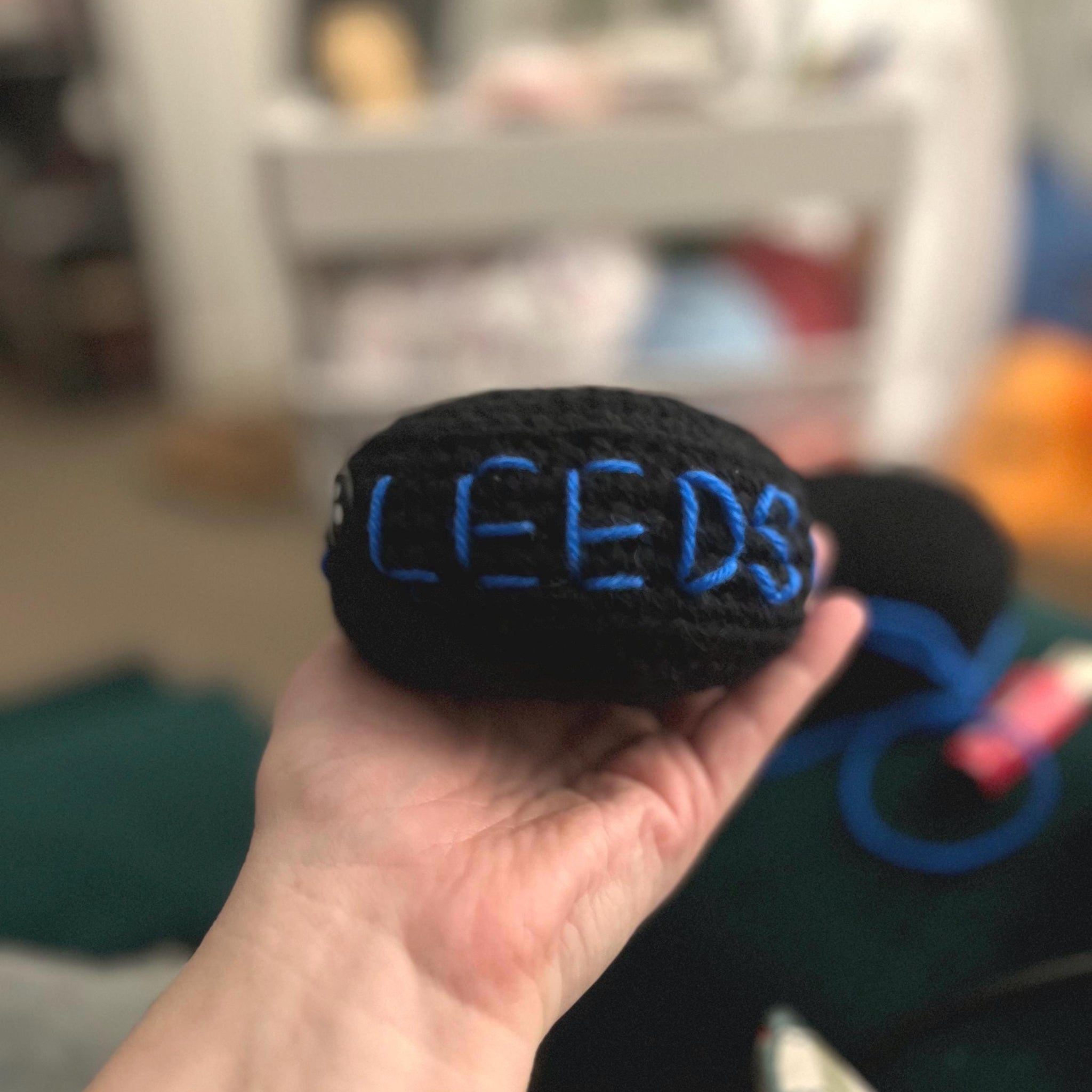 Hand holding one black crochet plush ice hockey puck with blue stitching of 'LEEDS'