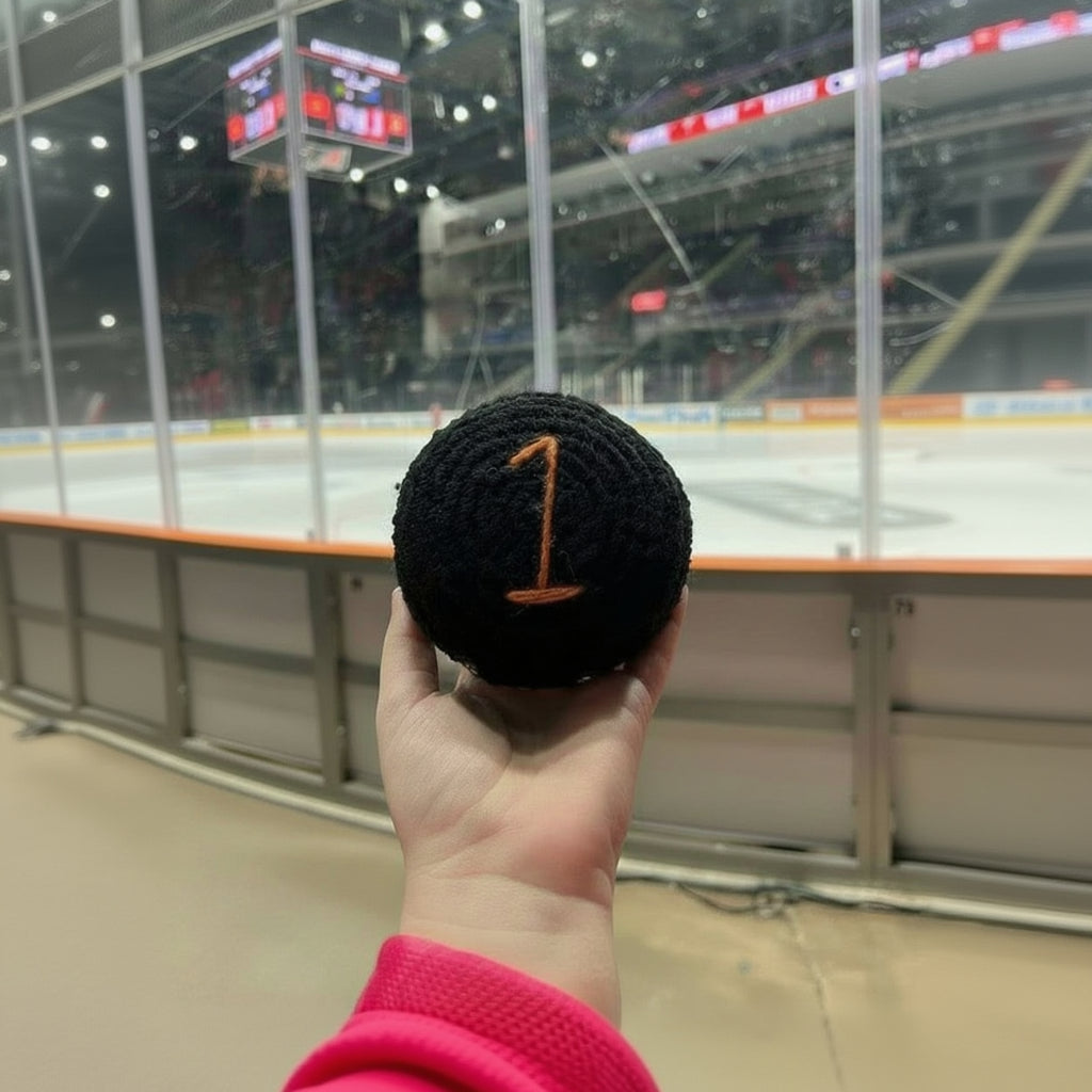 Hand holding a black crochet ice hockey puck with a number one on it in sheffield steelers orange accent, in front of an ice hockey rink.