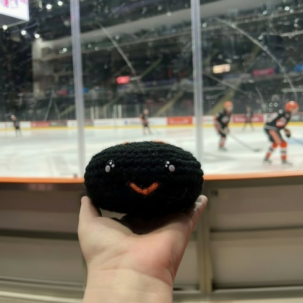 Hand holding a black crochet plush ice hockey puck with orange sheffield steelers colour themed accents in front of an ice hockey rink.