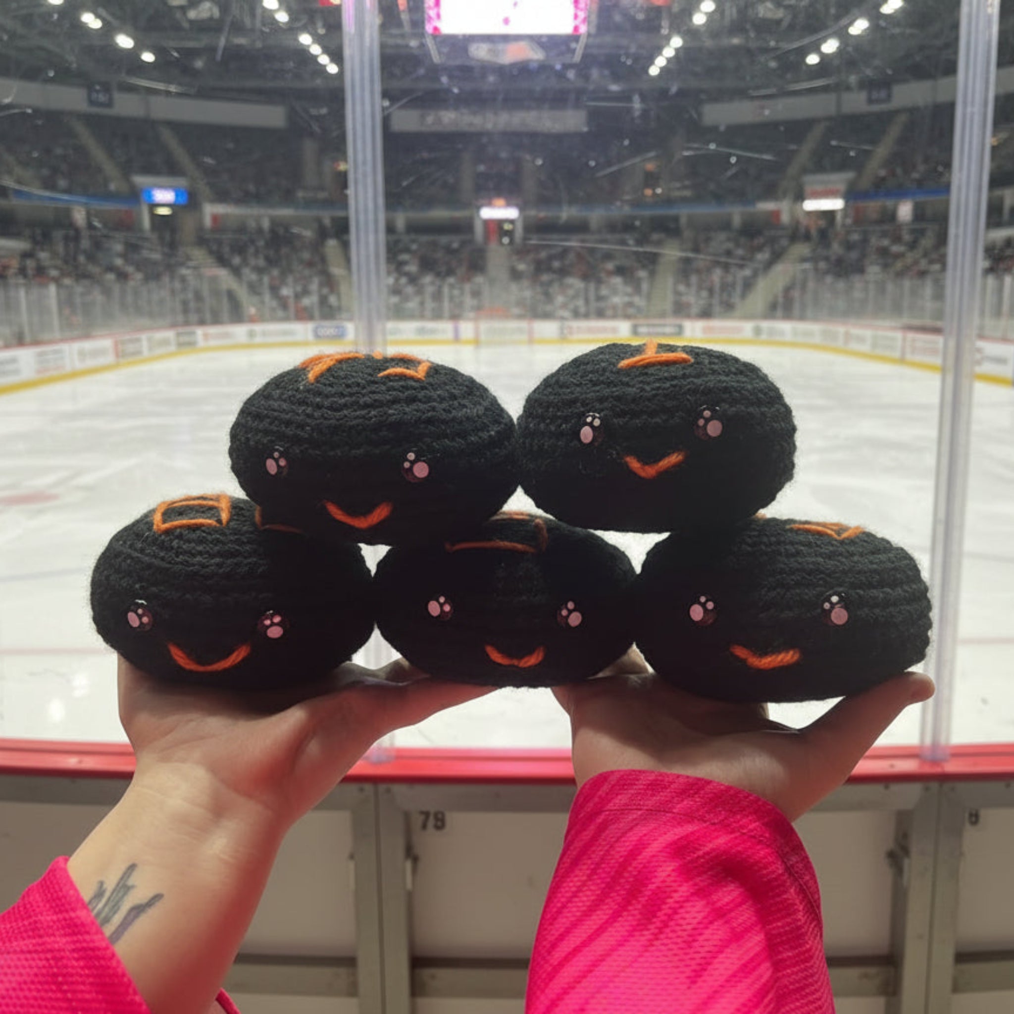 Hand holding five black crochet plush ice hockey puck toys with orange sheffield steelers colour themed accents in front of an ice hockey rink.