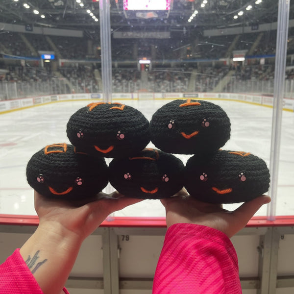 Hand holding five black crochet plush ice hockey puck toys with orange sheffield steelers colour themed accents in front of an ice hockey rink.
