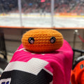 Person holding a sheffield steelers orange colour themed crochet ice hockey puck in front of an ice rink