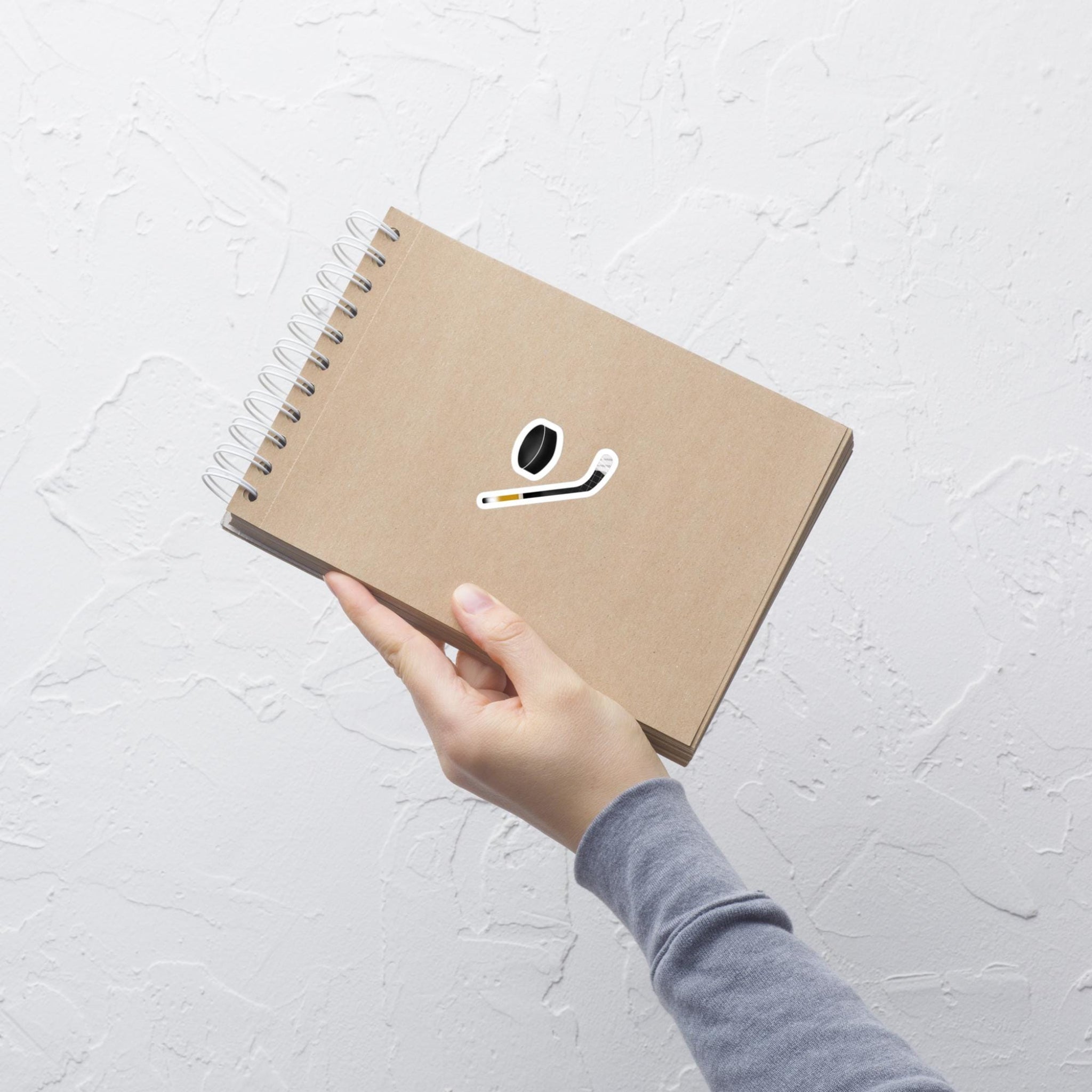 Hand holding brown notebook with a ice hockey sticker and ice hockey puck sticker in the center of the notebook